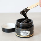 Black jar of Himalayan Shilajit with a wooden stick lifting some out, on a light background.