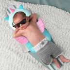 Baby lying on a colorful inflatable shark toy with sunglasses and gray shorts.