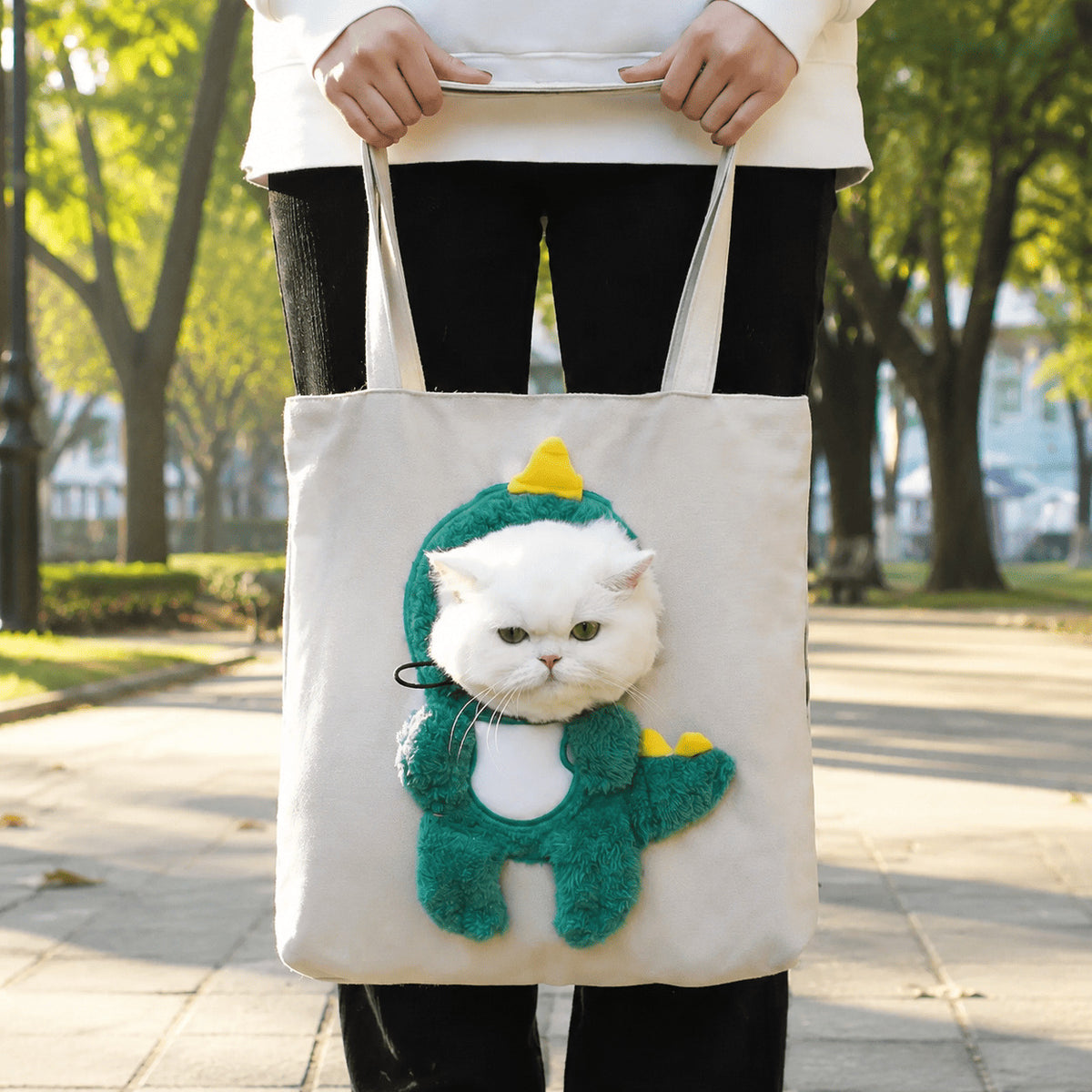 A person outdoors holding a cream canvas tote bag with a white cat dressed in a green dinosaur costume printed on the front.
