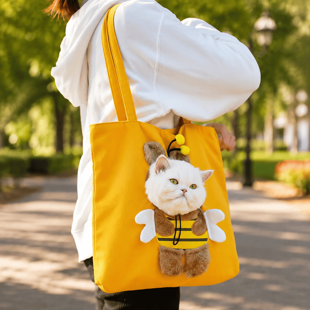 A person wearing a white hoodie carries a bright yellow tote bag on their shoulder in a sunny park. The bag features a plush white cat dressed in a yellow and black bee costume with white wings.