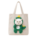 A beige canvas tote bag featuring a plush 3D applique of a white cat dressed in a green dinosaur costume with yellow accents, shown against a plain white background.